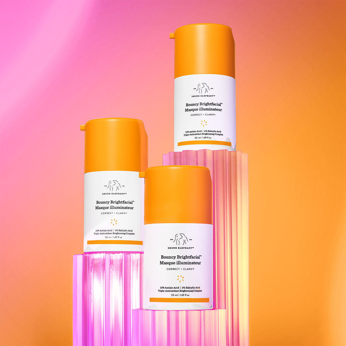 Mutiple Bouncy Brightfacial bottle on top of acrylic stands on a pink and orange background. 
