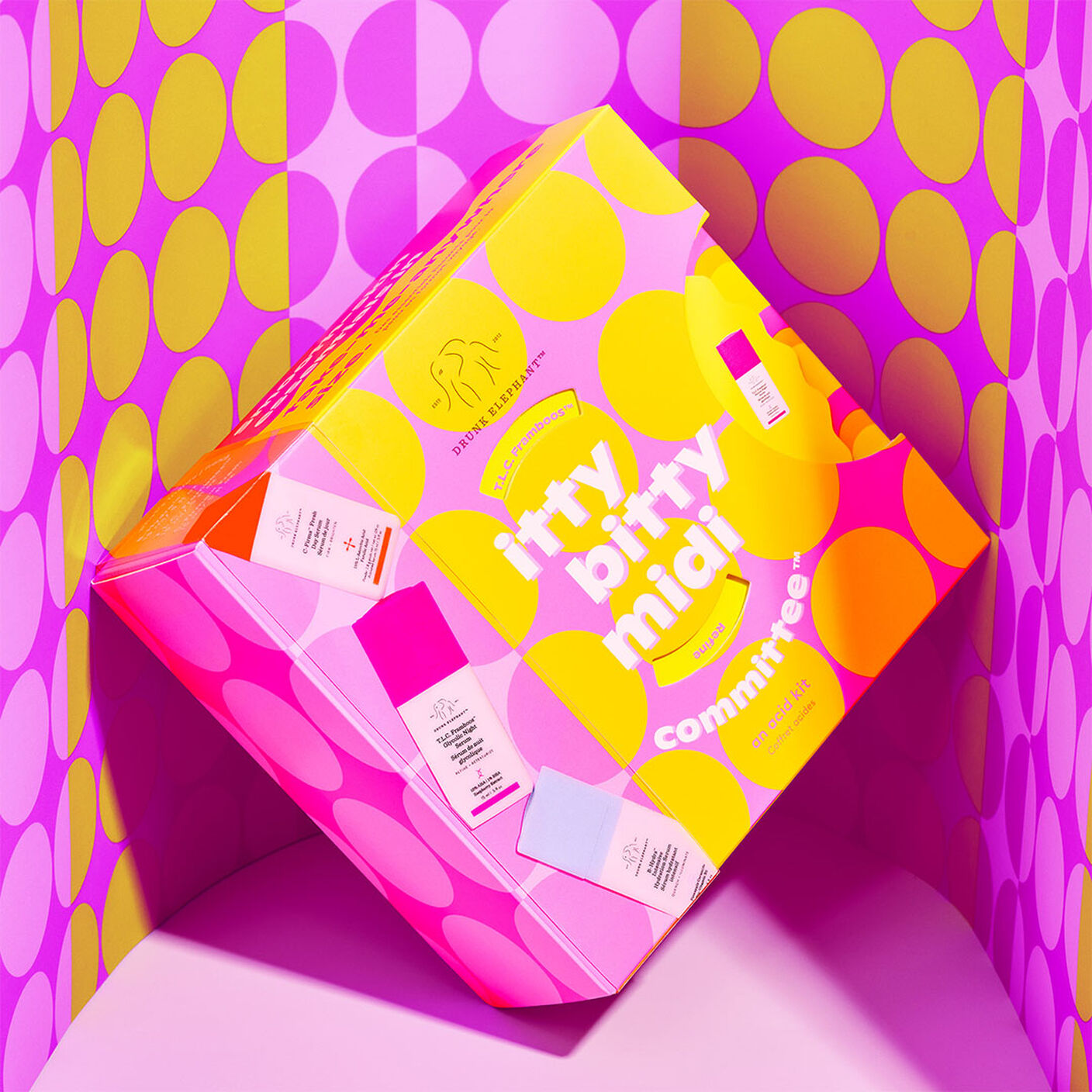 Image of packaging, a pink box with yellow circles, in front of a pink wall of yellow and pink circles. 