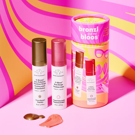 8ml of D-Bronzi and 8ml of O-Bloos next to pink and orange cylinder packaging on a pink floor in front of a pink and orange swirly background.
