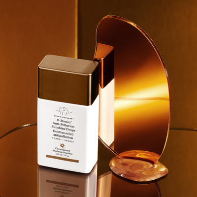 D-Bronzi™ Anti-Pollution Sunshine Drops against a brown stylized background and a mirror with product in front